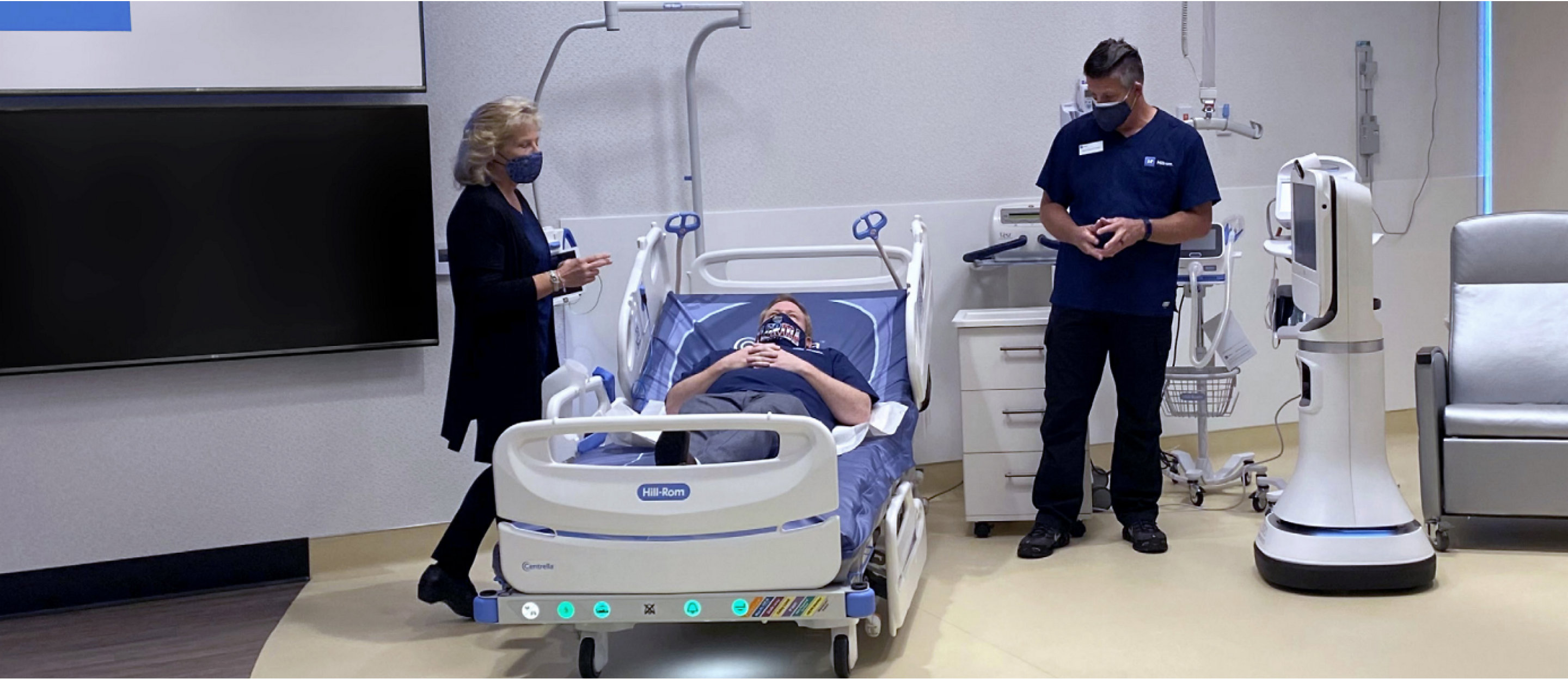 A patient smiles in a Centrella Smart+ Bed. Her clinician stands near the Graphical Caregiver Interface (GCI) Touchscreen.